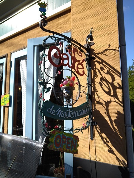 カフェドモトナミの看板。カラフルな金属製。店の名前が英語で書かれていて、真ん中にはフルーツパフェが飾ってある